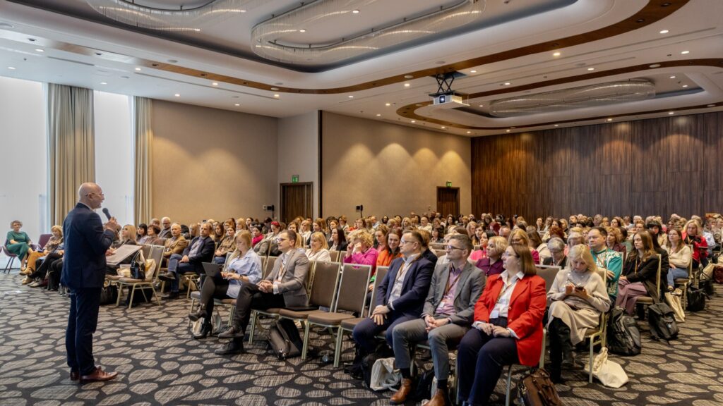 Zdjęcie przedstawia stojącego mężczyznę w garniturze (bokiem do obiektywu) przemawiającego do siedzących na dużej sali uczestników wydarzenia